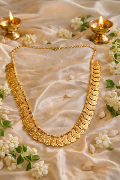 Lakshmi Haram Kasu Mala with traditional gold-tone coin detailing, laid flat on ivory satin with jasmine flowers and brass lamps.